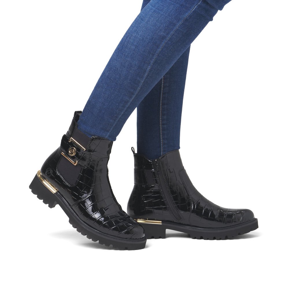 Lacquer black remonte women´s Chelsea boots D8684-03 with golden decorative element. Shoe on foot.